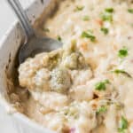 Cooked broccoli cauliflower casserole in a white baking dish on a white countertop. A serving spoon is scooping some of the casserole.