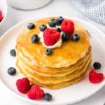 a stack of four fluffy homemade pancakes on a white plate with butter, syrup, blueberries and raspberries
