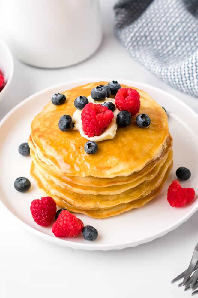 a stack of four fluffy homemade pancakes on a white plate with butter, syrup, blueberries and raspberries
