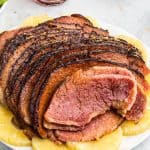 a cooked maple glazed ham sitting on a white serving platter with pineapple around the edges