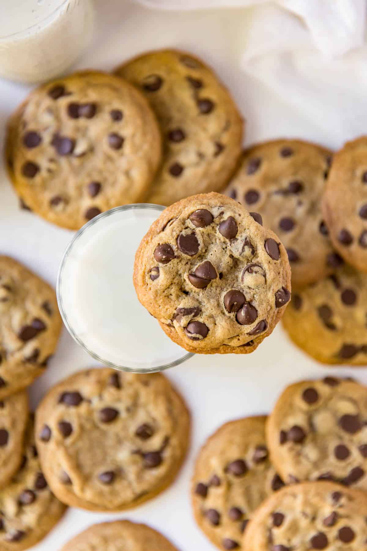 a chocolate chip cookie sitting on a cup of milk with a pile of cookies around the glass