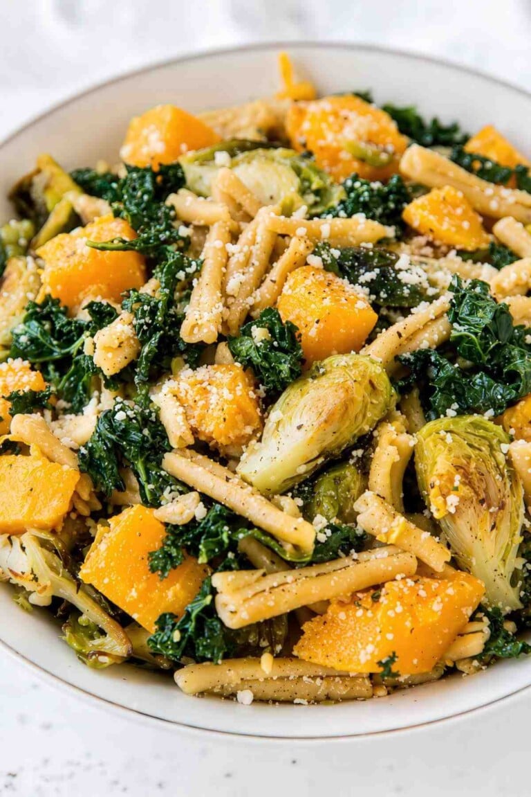 Close-up of chickpea pasta with Brussels sprouts, butternut squash, kale, and grated Parmesan in a white bowl.