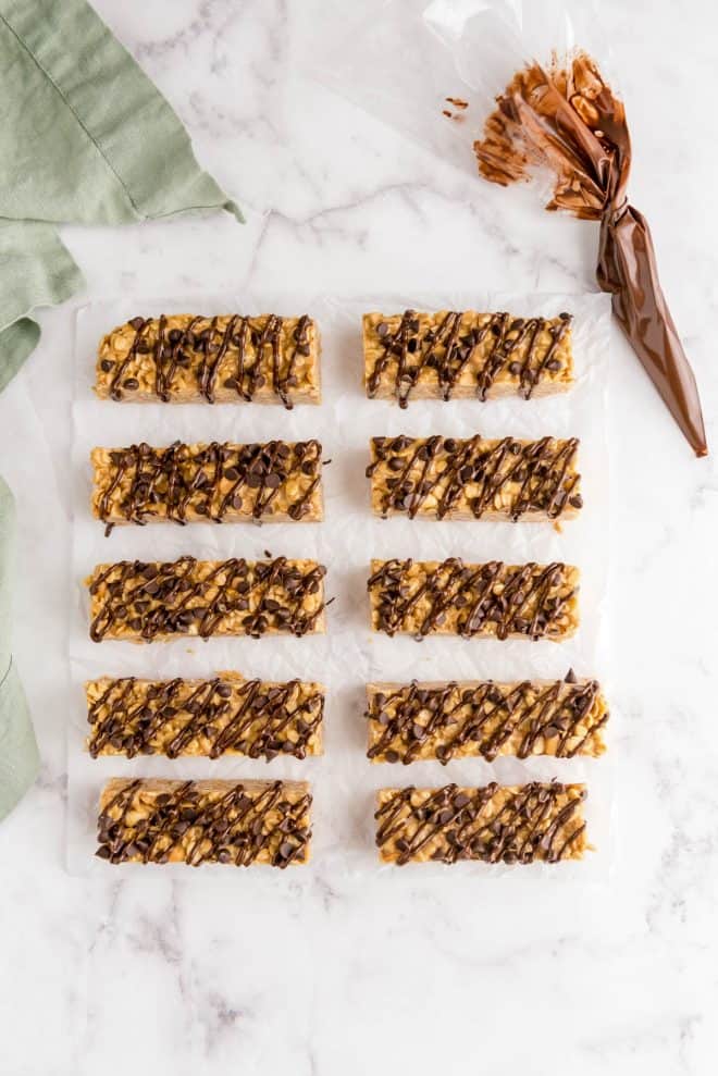 chocolate peanut butter granola bars sitting on white parchment paper with a piping bag filled with chocolate on the side