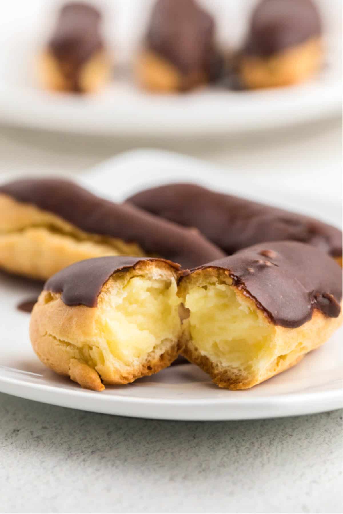 Three chocolate eclairs on a plate, one cut in half to show the vanilla cream filling. 