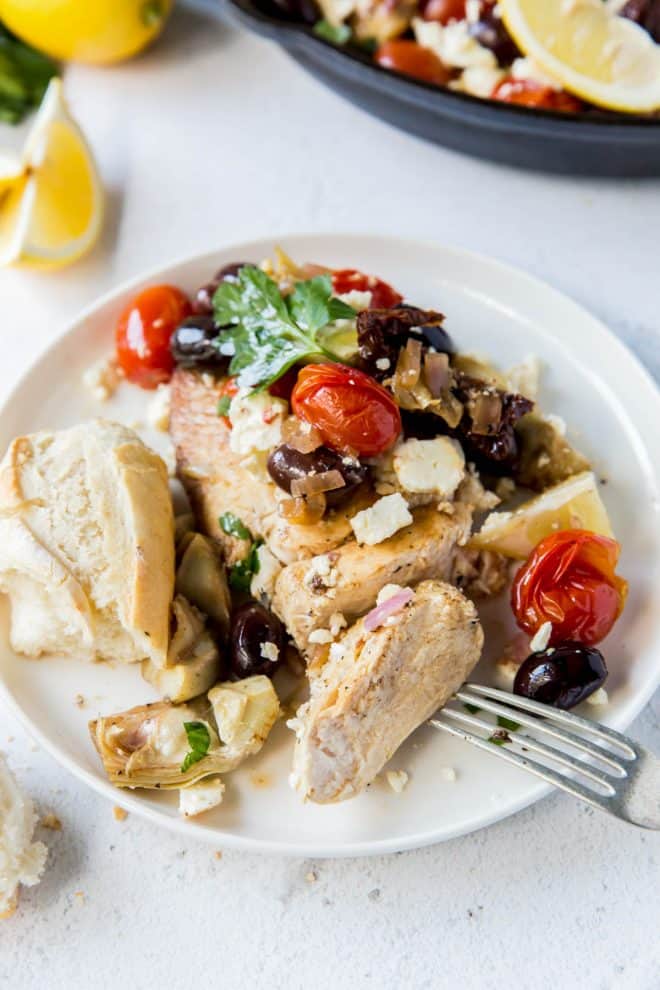 a piece of chicken on a plate with tomatoes, olives, feta and artichokes on top