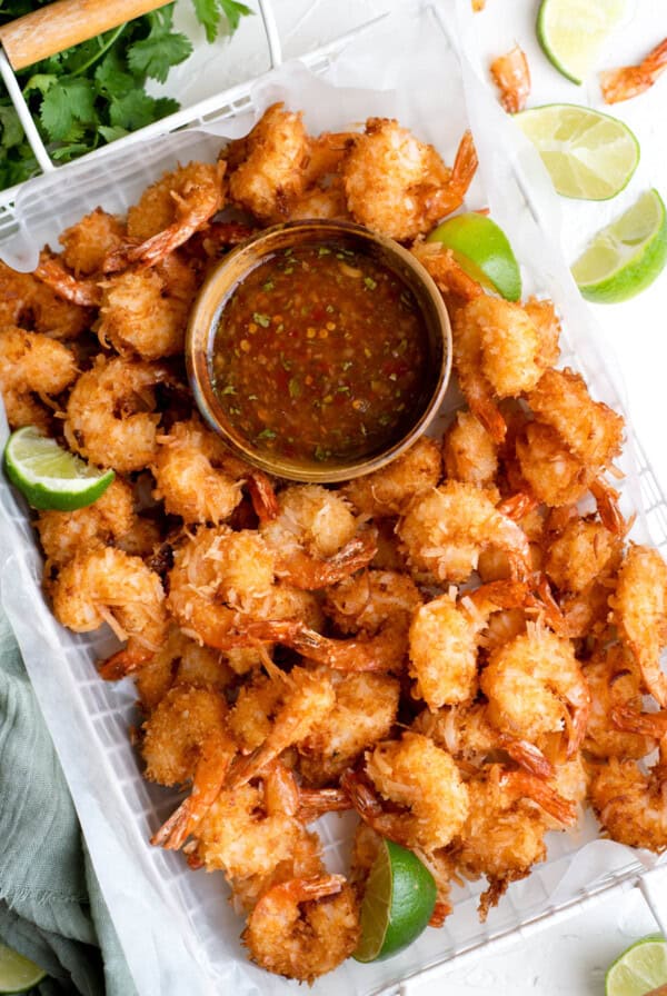 Golden, crispy coconut-coated shrimp arranged around a bowl of sweet chili lime dipping sauce, served with fresh lime wedges on a parchment-lined tray.
