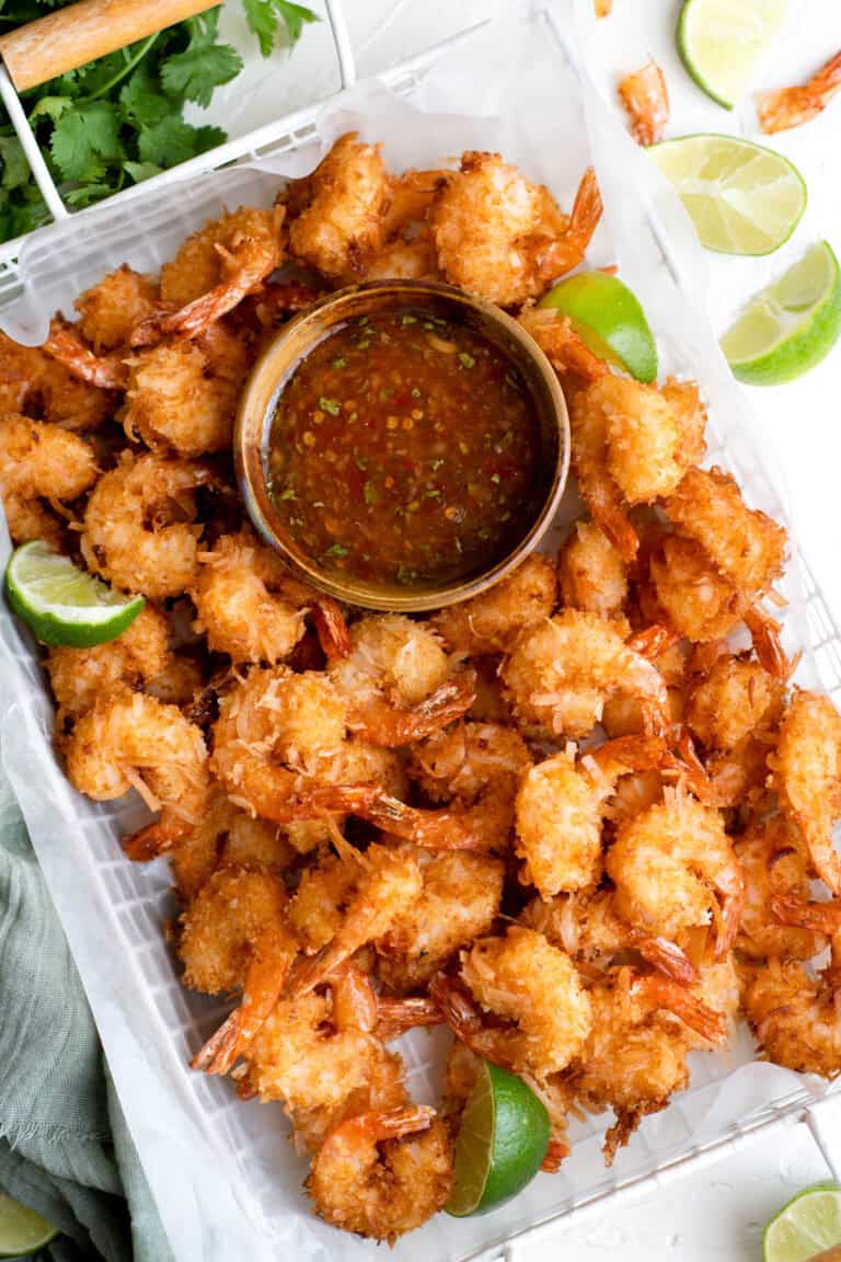 Golden, crispy coconut-coated shrimp arranged around a bowl of sweet chili lime dipping sauce, served with fresh lime wedges on a parchment-lined tray.