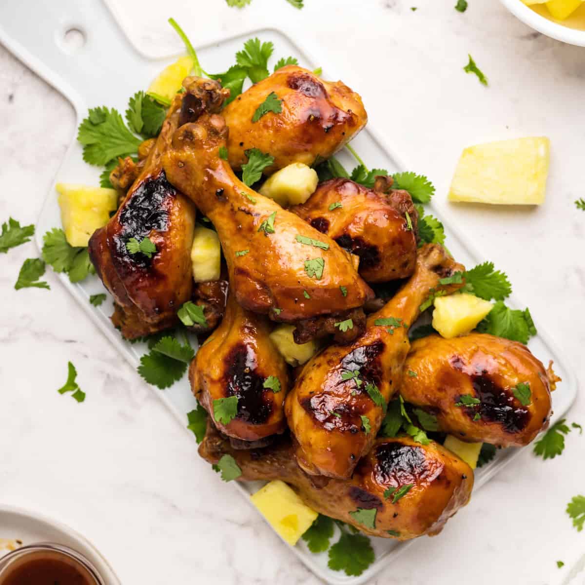Overhead view of Hawaiian BBQ chicken drumsticks served on a white platter with fresh pineapple chunks and cilantro, surrounded by dipping sauce, tongs, and a bowl of pineapple pieces.