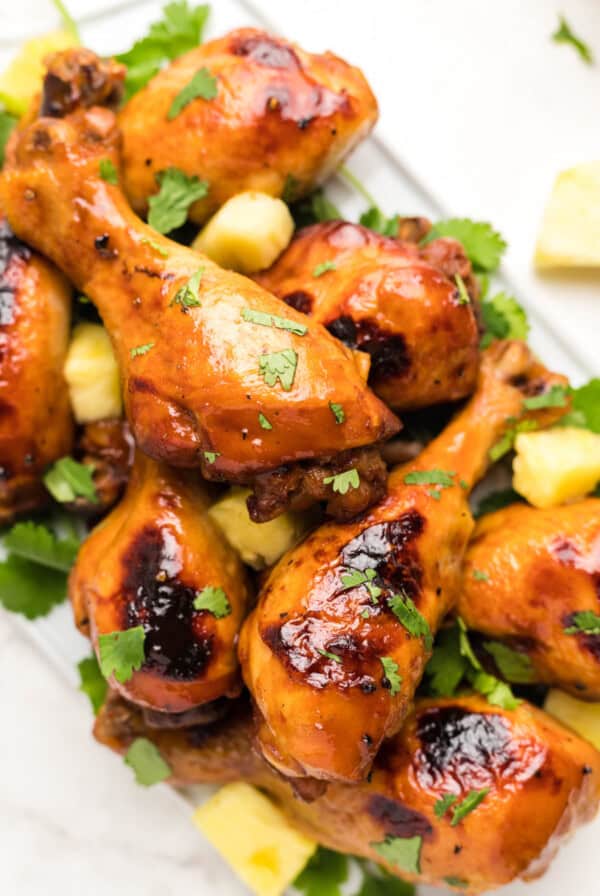 Close-up overhead view of Hawaiian BBQ chicken drumsticks garnished with chopped cilantro and pineapple chunks on a white platter