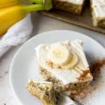 A banana bar with cream cheese icing on a plate, with bananas and more bars in the background.