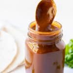 A glass jar of homemade enchilada sauce with a spoon stirring the sauce.