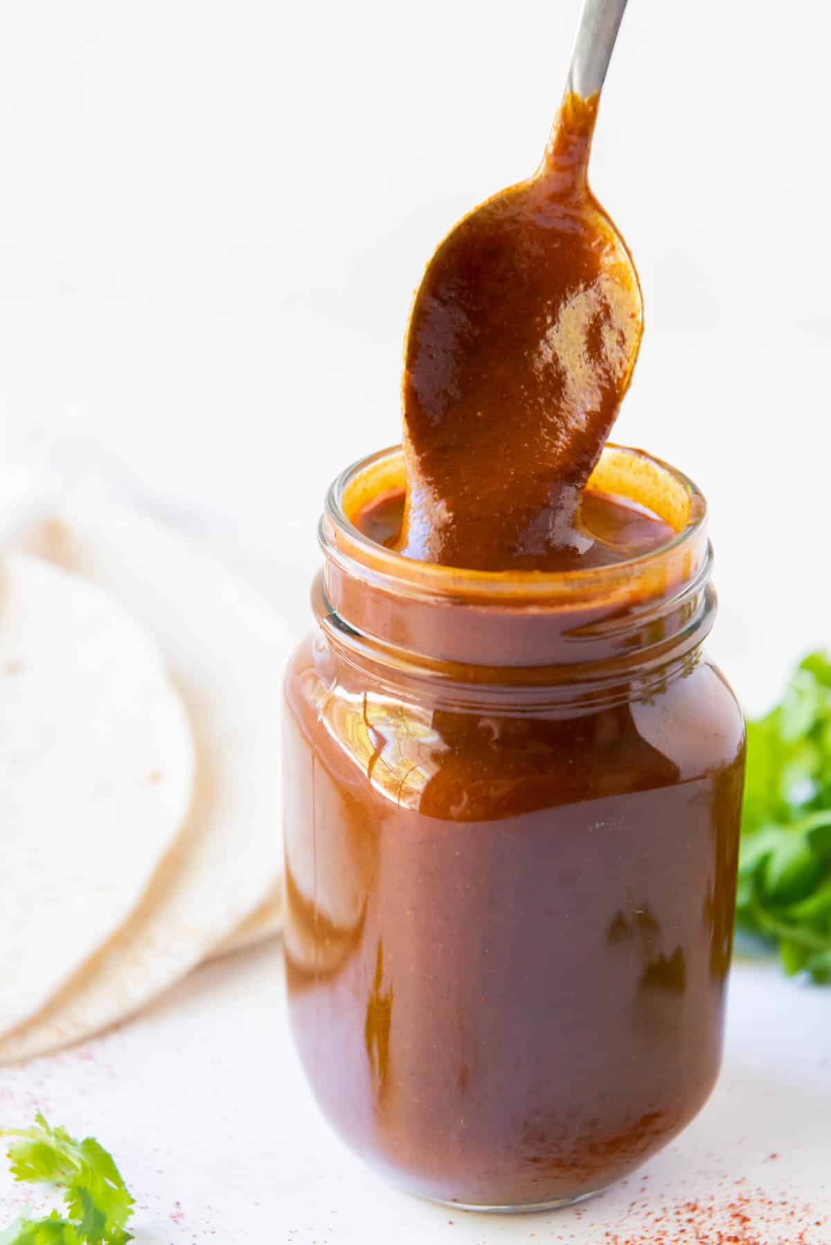 A glass jar of homemade enchilada sauce with a spoon stirring the sauce.