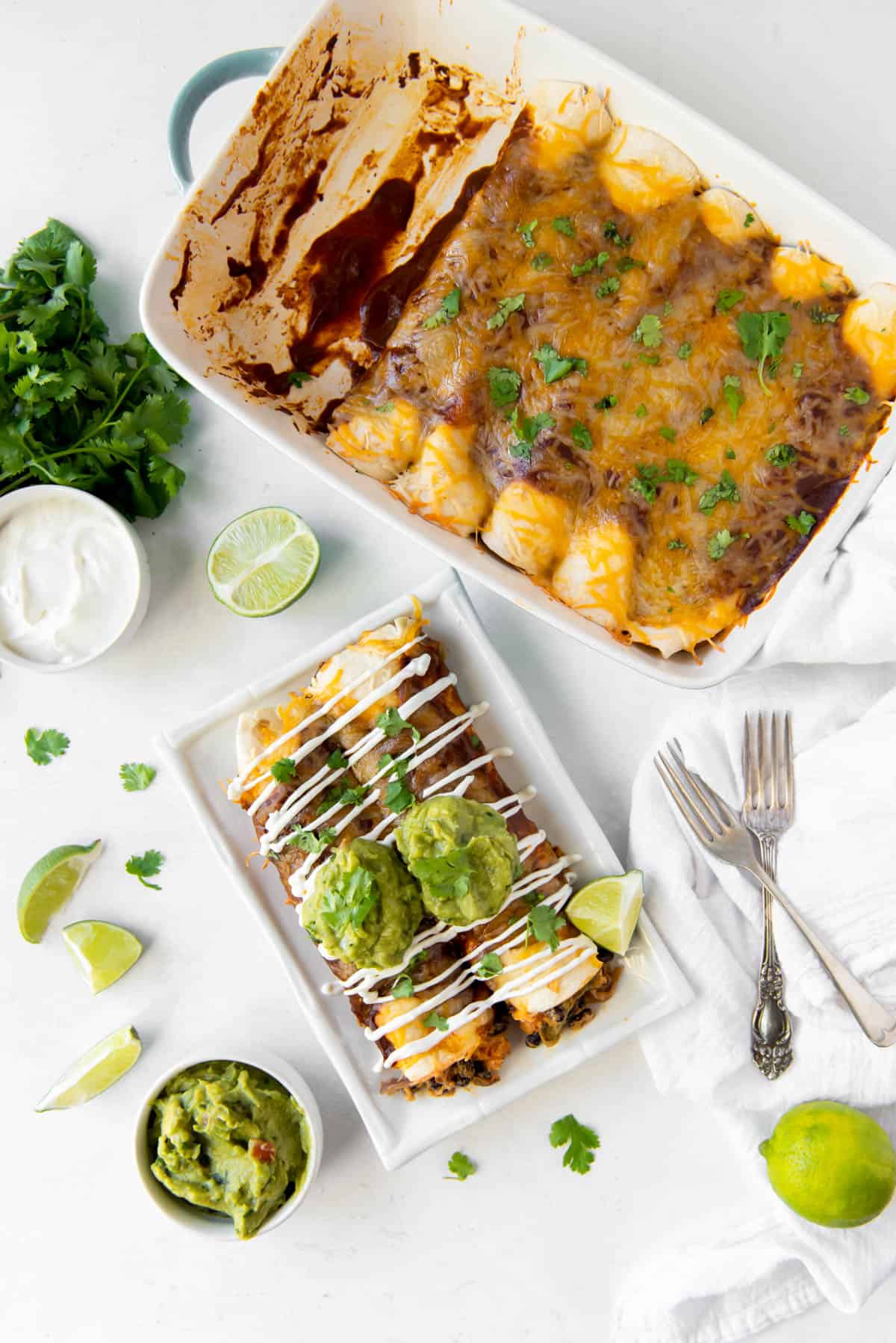 Aerial view of a pan of veggie enchiladas and a plate with two vegetable enchiladas, topped with guacamole and sour cream. 