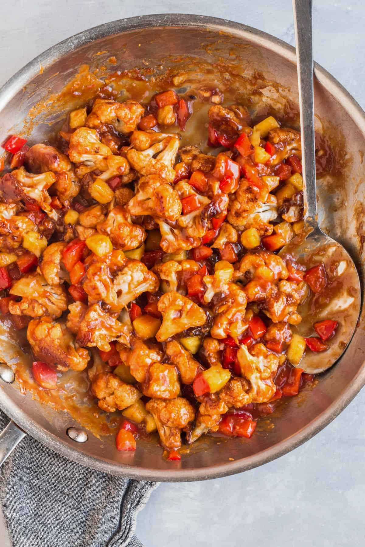 Sweet and sour cauliflower tossed with pineapple and red bell pepper in a skillet, coated in a thick, glossy sauce.