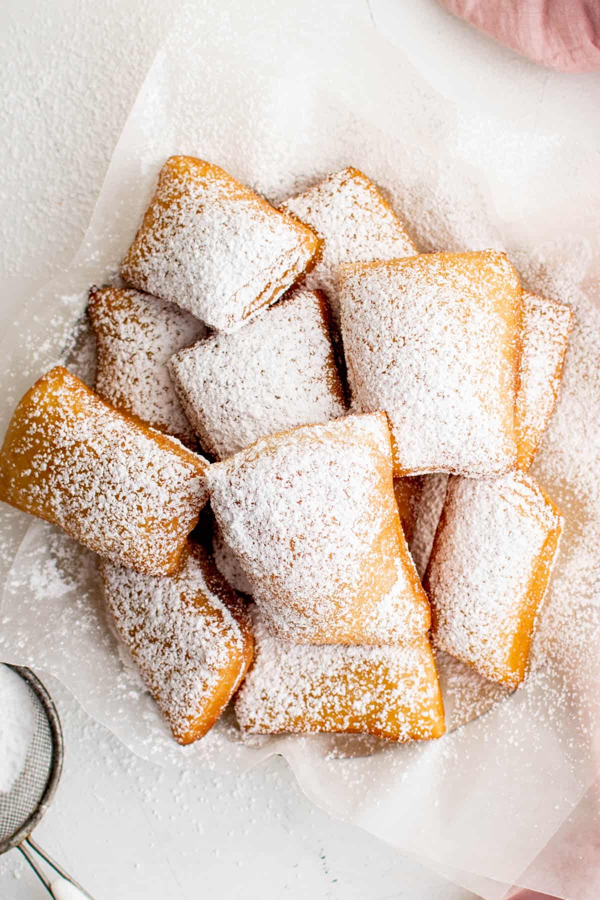 A large pile of sugar dusted beignets sitting on a piece of parchment paper.