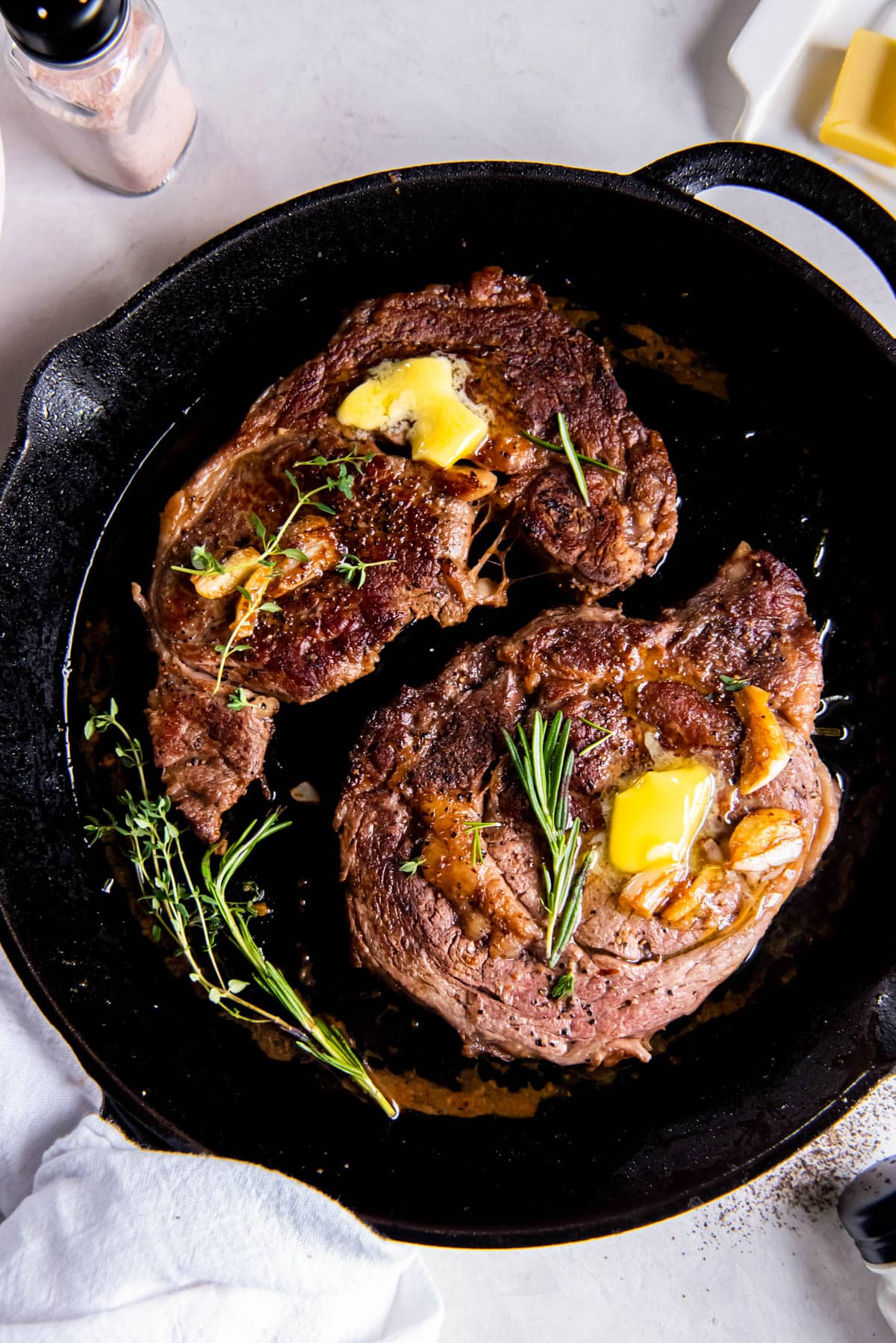 Two pan seared ribeye steaks in a cast iron skillet with a slice of melted butter and fresh rosemary on top.