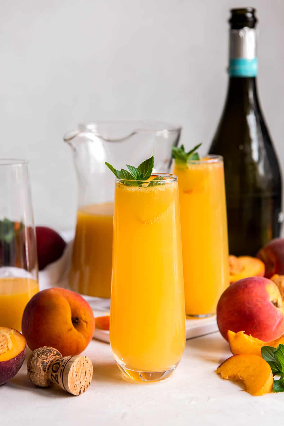 Two glasses of fresh peach mimosas sitting on a white counter with a bottle of prosecco in the background.
