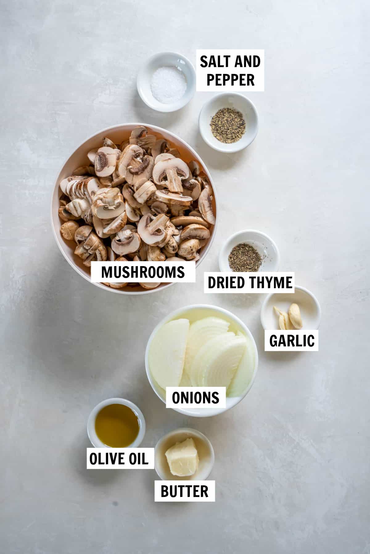 All of the ingredients for sauteed mushrooms and onions on a white countertop including mushrooms, onion, garlic, olive oil, salt, pepper and thyme.