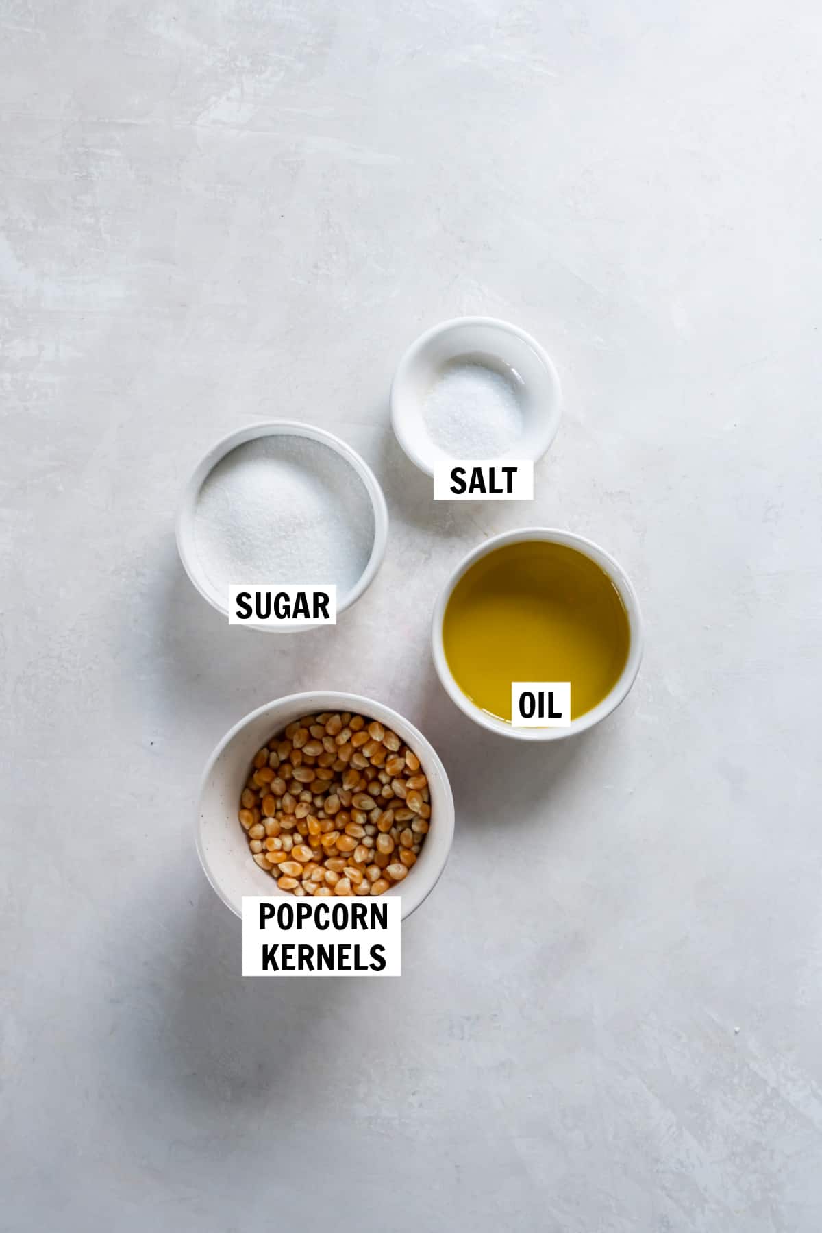 All of the ingredients for sweet and salty popcorn on a white countertop including popcorn kernels, oil, sugar and salt.