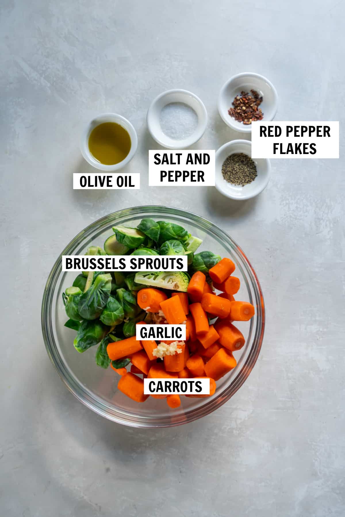 All of the ingredients for roasted brussels sprouts and carrots in bowls on a white countertop including fresh brussels sprouts, fresh cut carrots, minced garlic, olive oil, sea salt, black pepper and red pepper flakes.