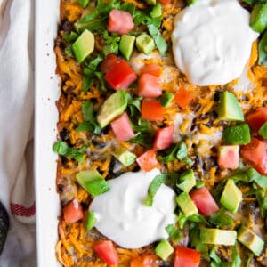A white casserole dish filled with taco casserole and topped with chopped tomato, avocado, cilantro and sour cream.