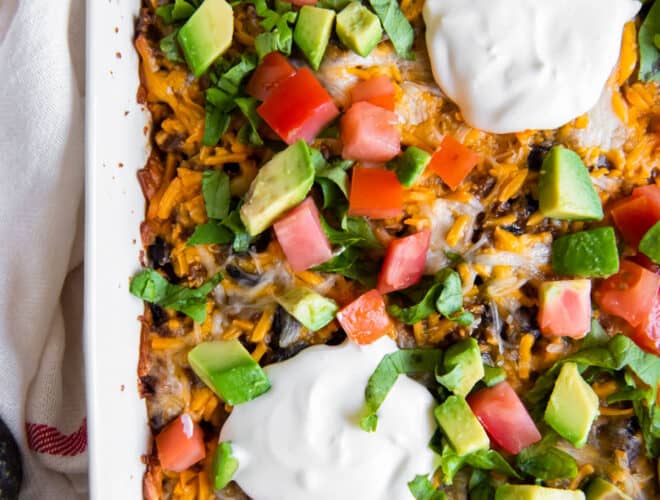 A white casserole dish filled with taco casserole and topped with chopped tomato, avocado, cilantro and sour cream.