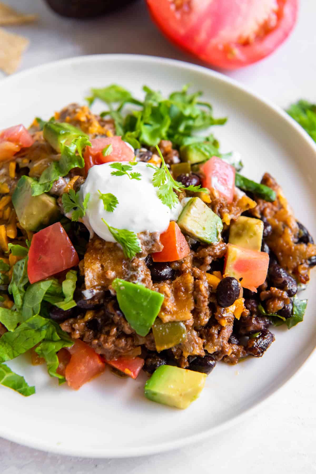 A scoop of taco casserole on a white plate topped with fresh avocado, tomato, sour cream and cilantro. 