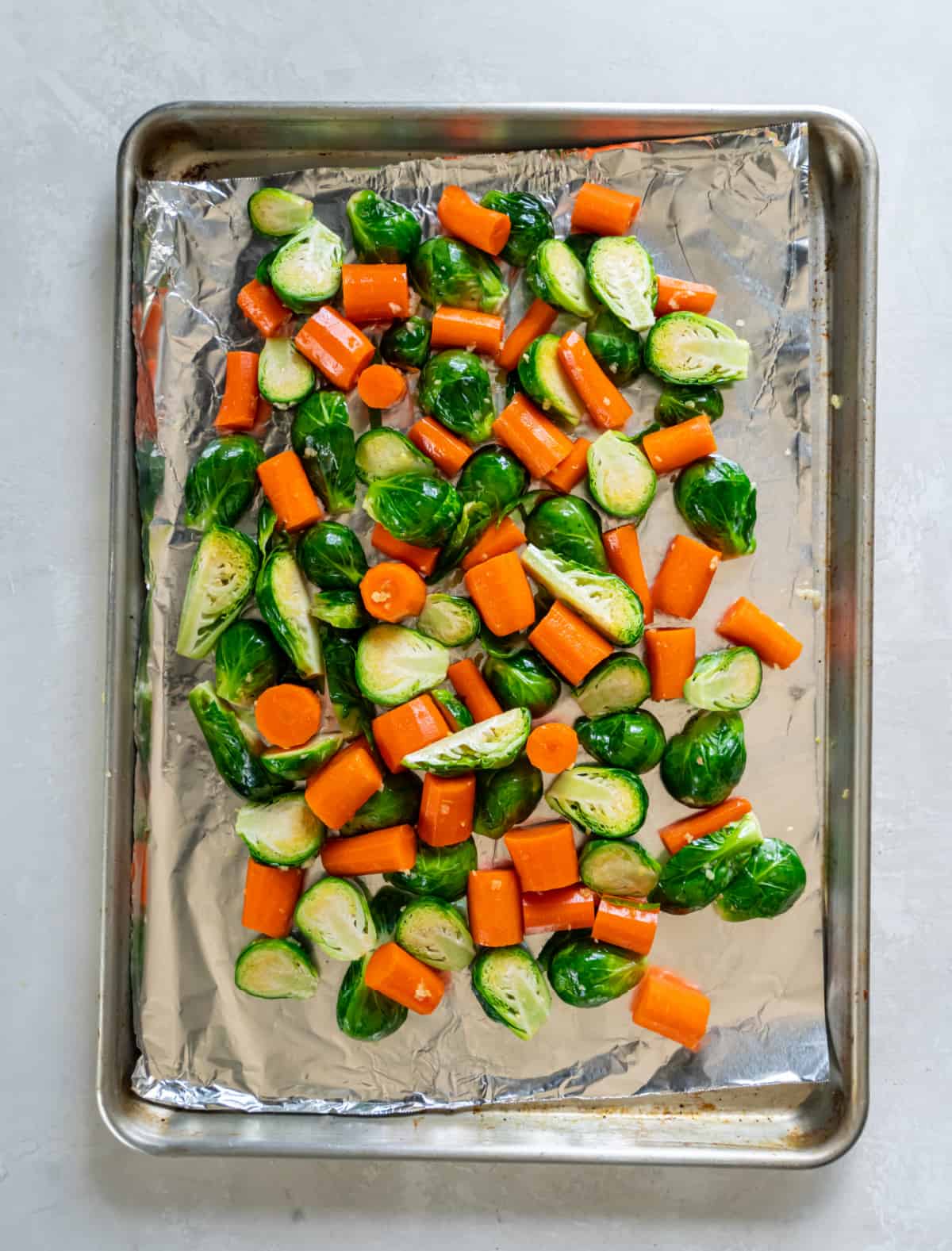 Brussels sprouts and carrots on a sheet pan with olive oil, garlic, salt, pepper, and red pepper flakes. The pan is sitting on a white countertop before going into the oven.