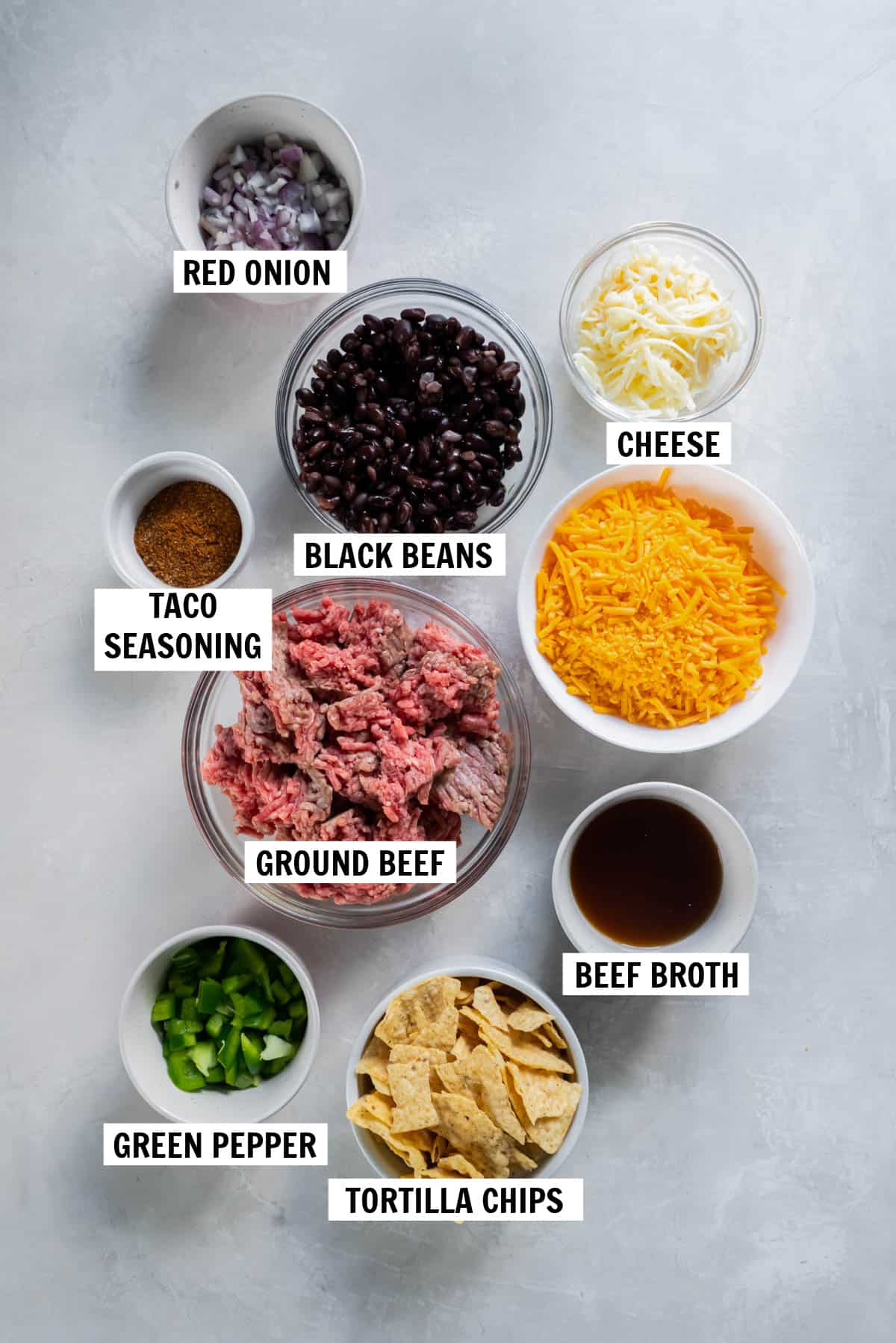 All of the ingredients for taco casserole including ground beef, black beans, cheese, onions, taco seasoning, bell pepper, beef broth and tortilla chips.