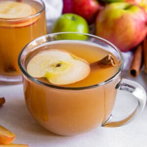 Two glasses of apple cider sitting on a white countertop. A slice of apple and one cinnamon stick are in the glass of cider.