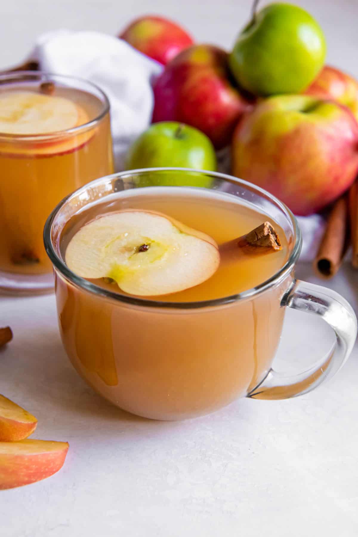Two glasses of apple cider sitting on a white countertop. A slice of apple and a cinnamon stick are in the cider as a garnish.