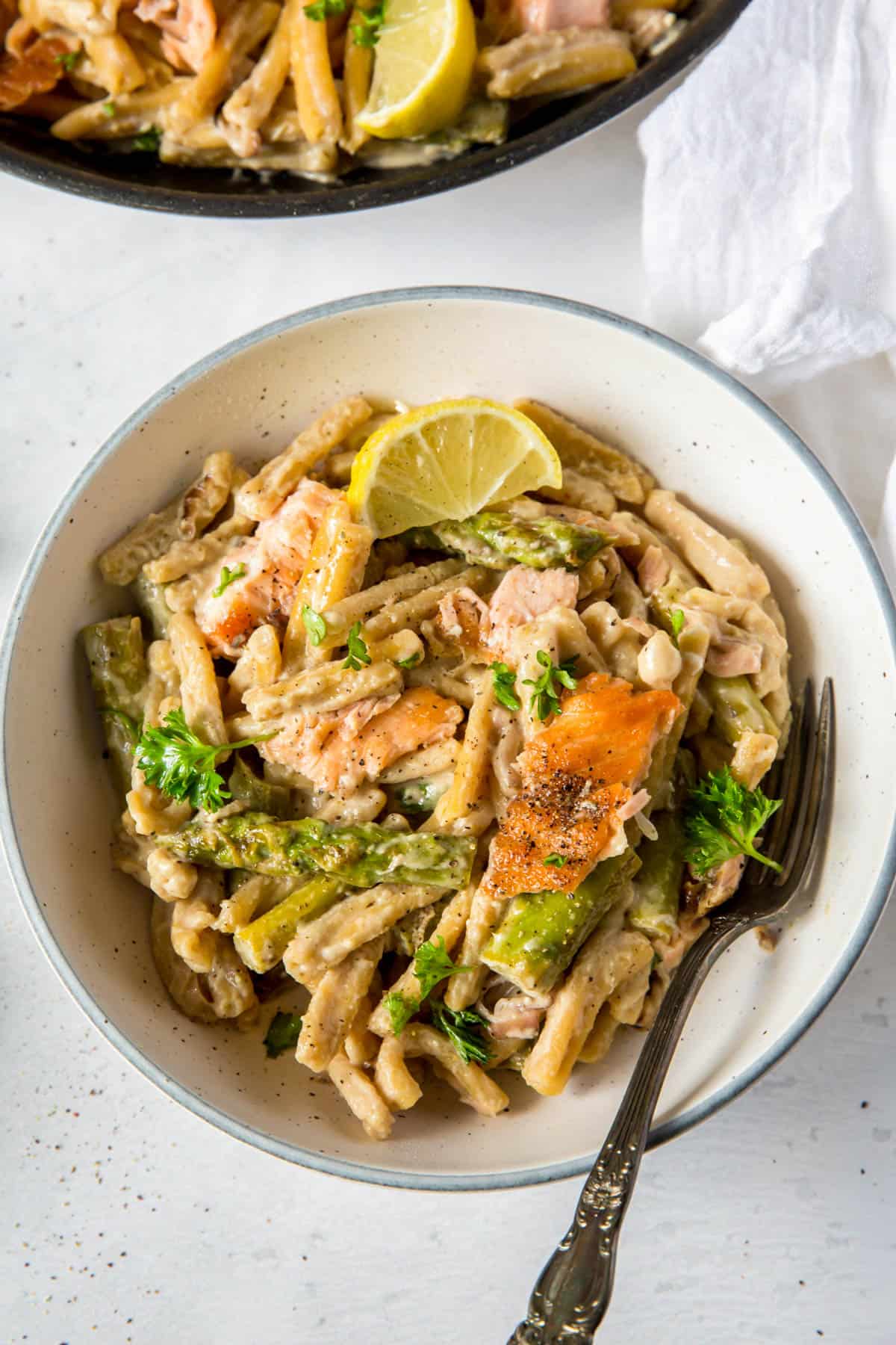 A white bowl filled with cooked creamy salmon pasta. A fork rests in the bowl and a lemon wedge is sitting on the top of the pasta.