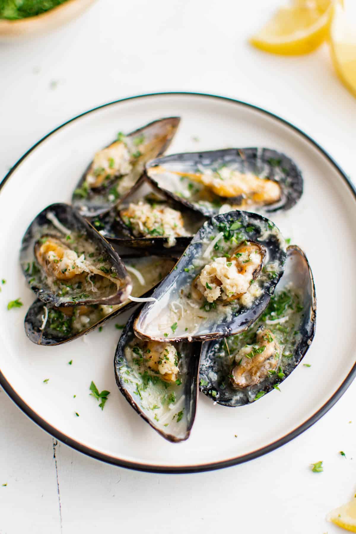 Eight baked mussels in garlic butter on a white plate sitting on a white tabletop for serving. They are garnished with freshly chopped parsley.