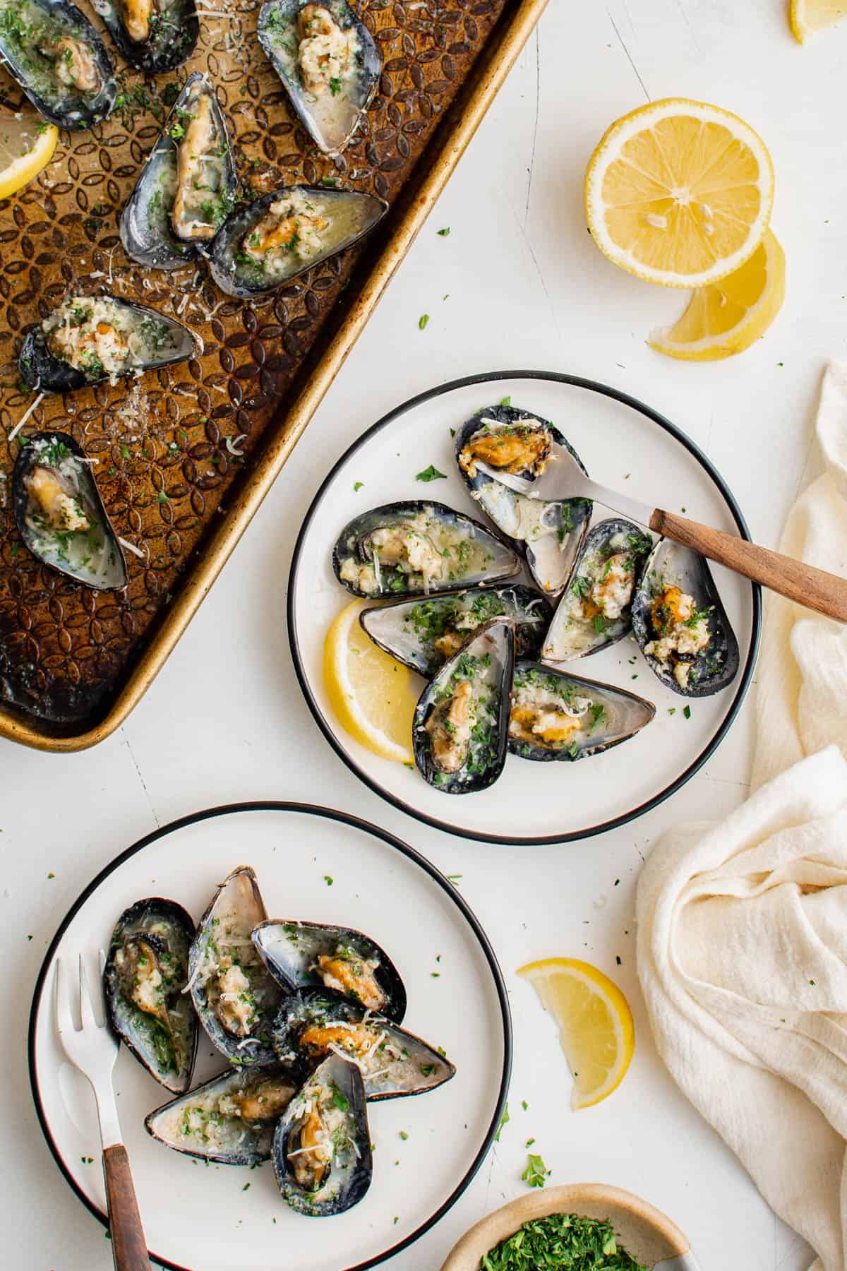Baked mussels in garlic butter arranged on white plates for serving with lemon wedges.