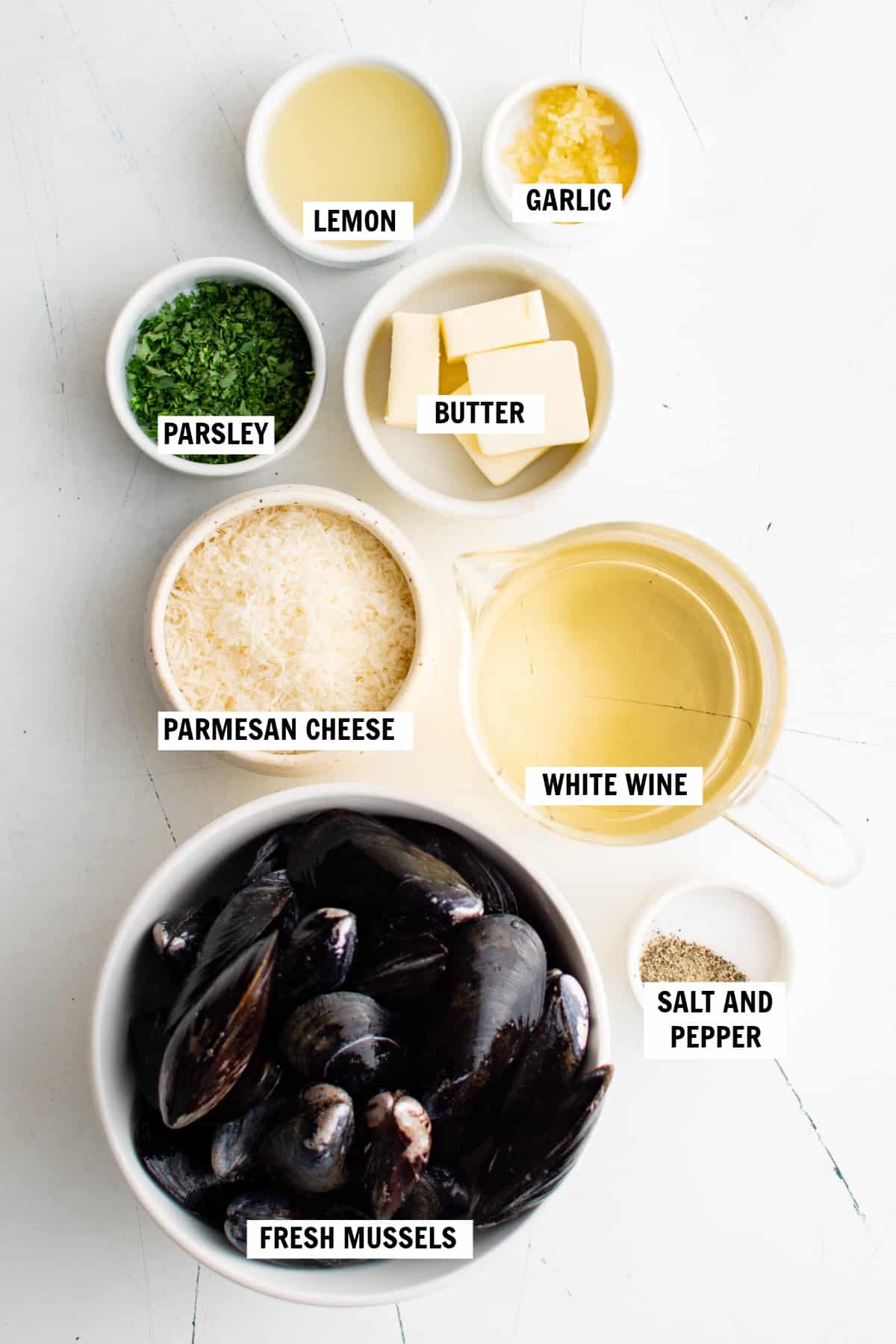 All of the ingredients for baked mussels on a white countertop including white wine, lemon juice, fresh mussels, butter, garlic, salt, pepper, Parmesan cheese and minced parsley.
