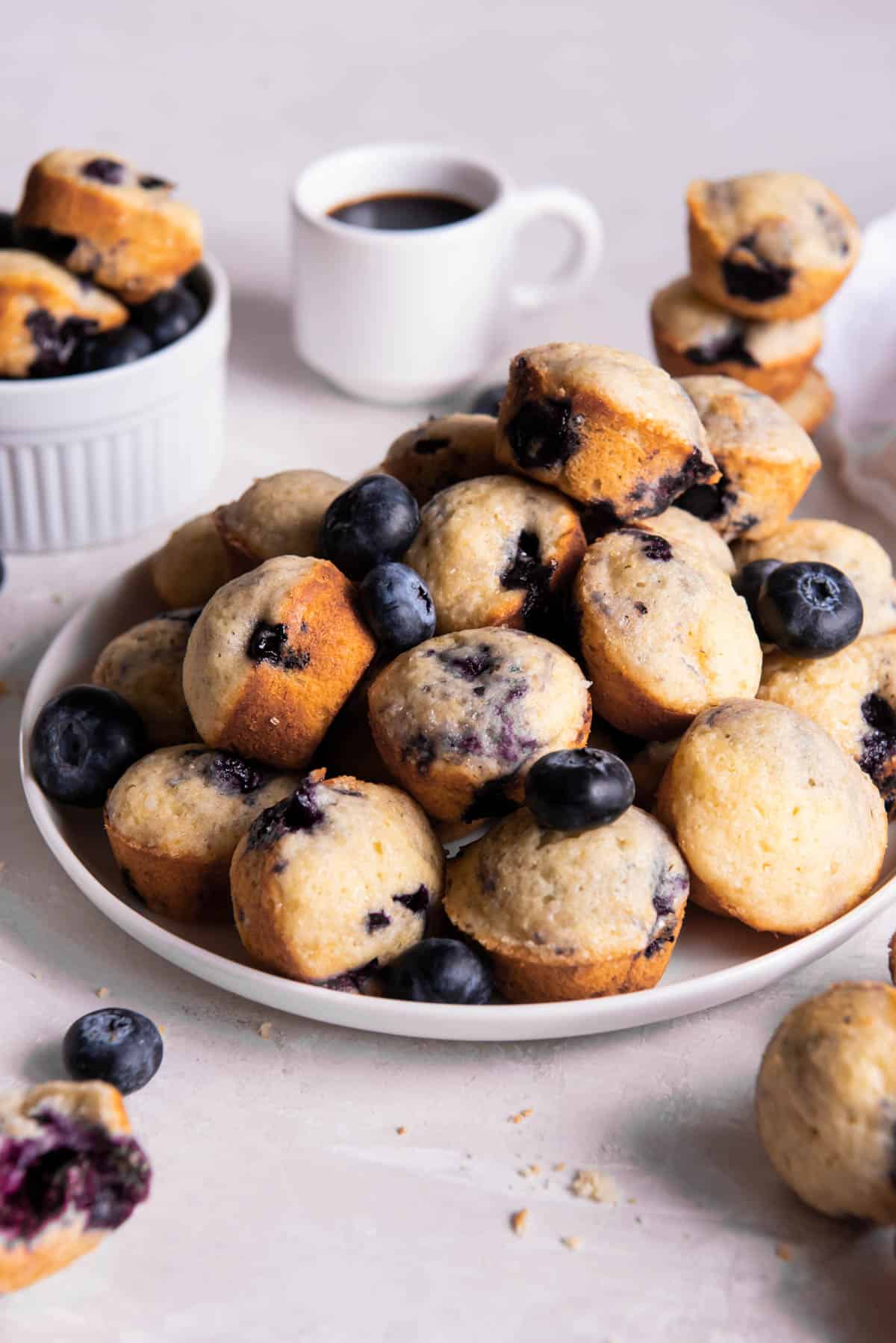 A large stack of mini blueberry muffins sitting on a white plate on a countertop. A cup of coffee sits in the background.
