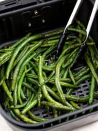 Tongs serving cooked air fryer green beans out of the air fryer basket.