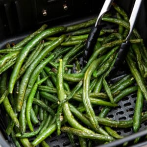 Tongs serving cooked air fryer green beans out of the air fryer basket.