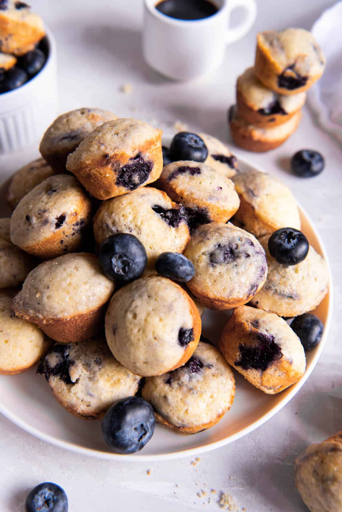 Mini blueberry muffins piled high on a white plate. Fresh blueberries are scattered on top of the mini muffins for serving.