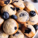 Mini blueberry muffins sitting on a plate for serving. Fresh blueberries are scattered on top of the muffins.