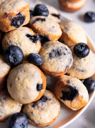 Mini blueberry muffins sitting on a plate for serving. Fresh blueberries are scattered on top of the muffins.