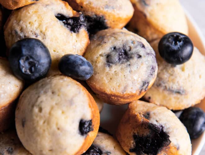 Mini blueberry muffins sitting on a plate for serving. Fresh blueberries are scattered on top of the muffins.