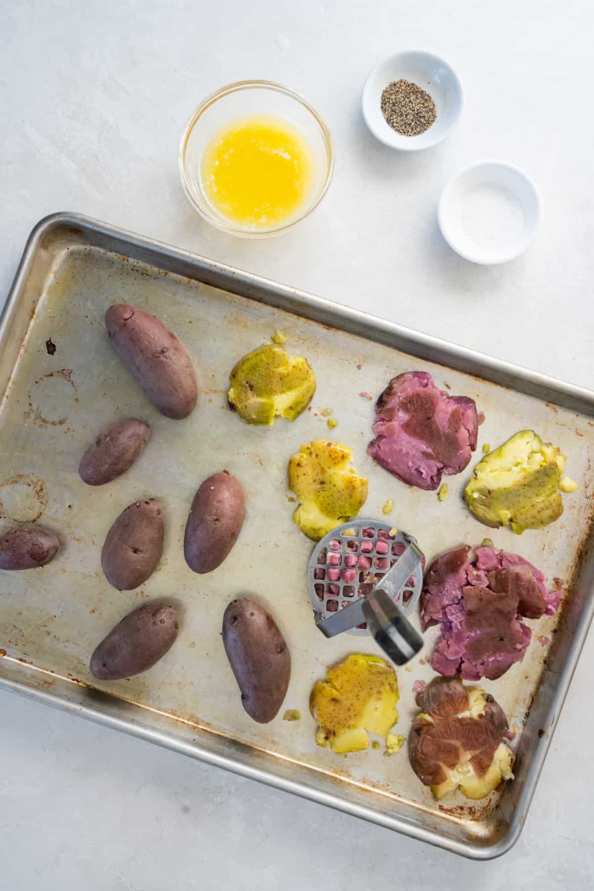 Fingerling potatoes sitting on a sheet pan with a potato mashed pressed down on one of the potatoes to smash it.