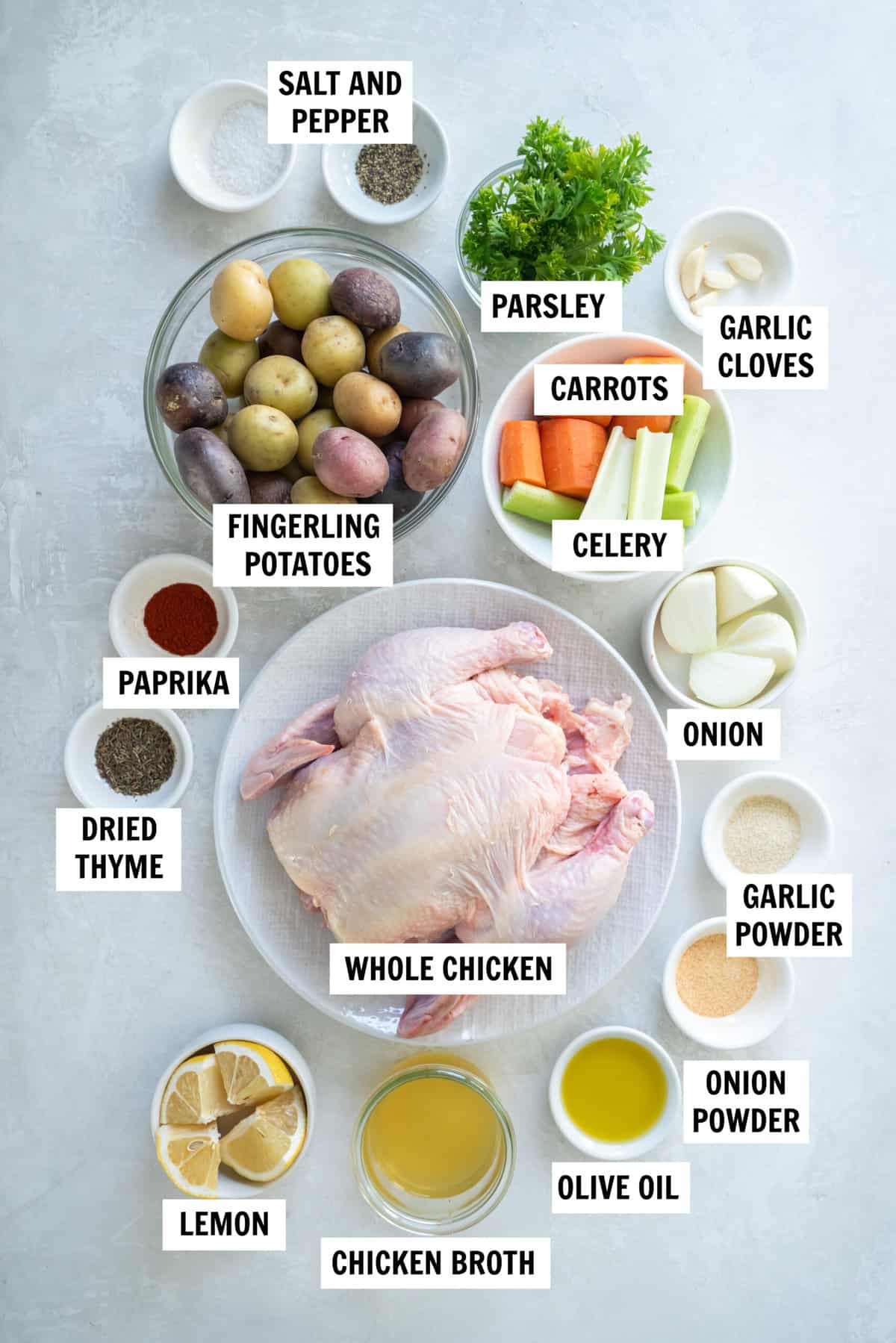 All of the ingredients for dutch oven whole chicken on a white countertop including whole chicken, celery, carrots, lemon, onion, chicken broth, olive oil, finergling potatoes, garlic and spices.