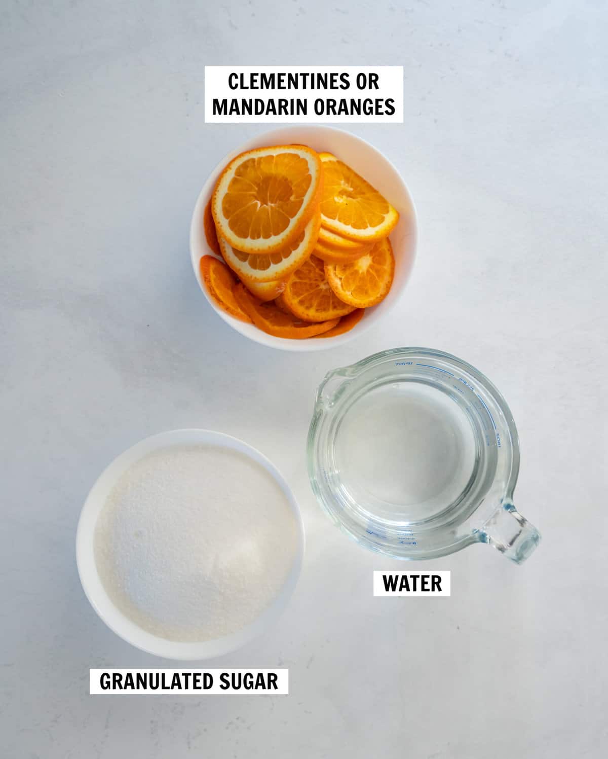 All of the ingredients for candied orange peels on a white countertop including orange slices, granulated sugar and water.