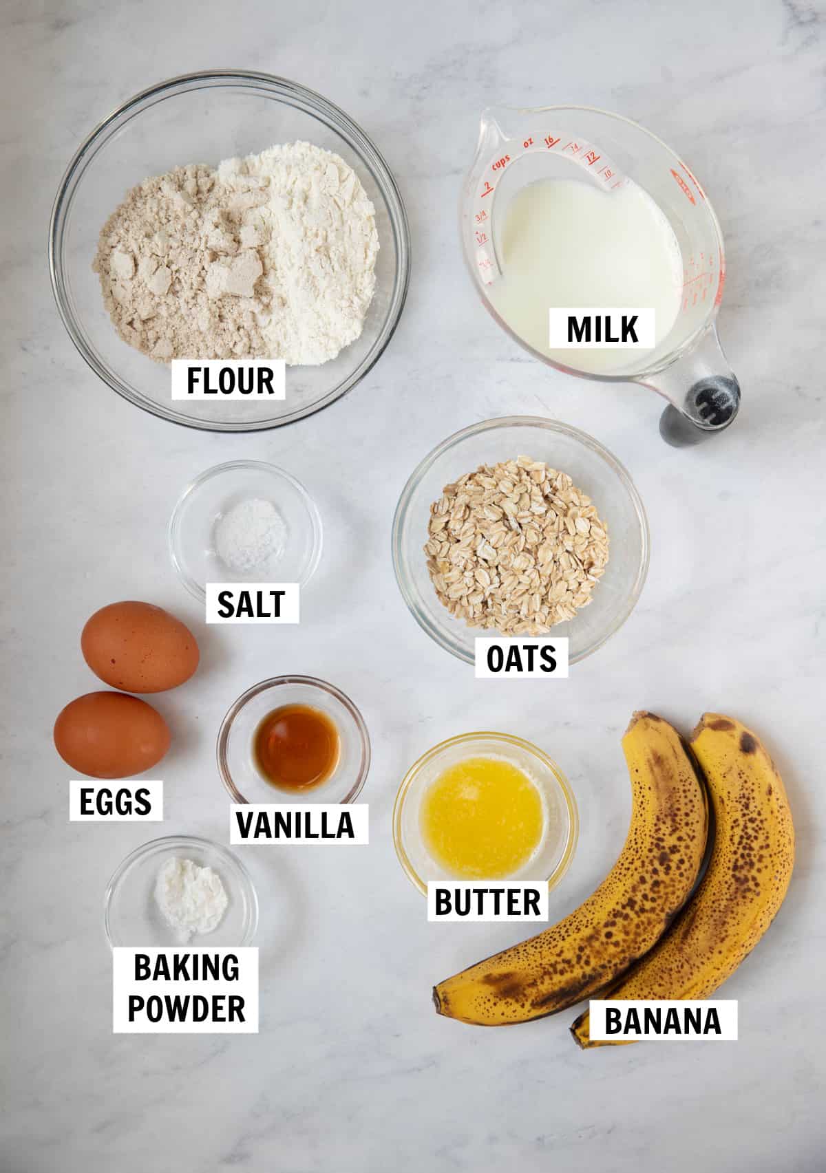 All of the ingredients for banana oat pancakes on a white countertop including oat flour, all purpose flour, oats, salt, baking powder, eggs, melted butter, banana, milk and vanilla extract.