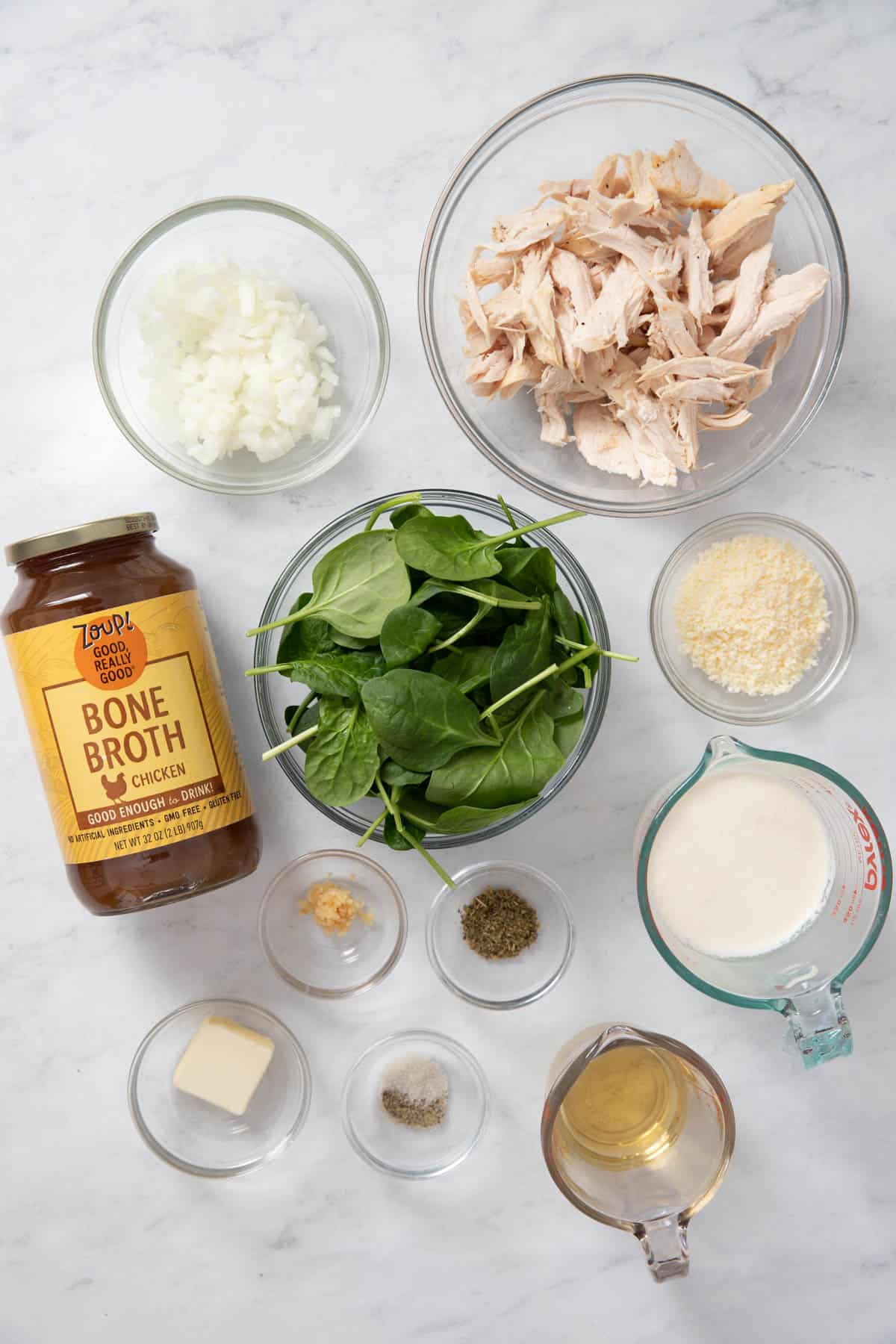 All of the ingredients for chicken florentine soup on a white countertop including butter, onion, garlic, chicken bone broth, heavy cream, white wine, Italian seasoning, rotisserie chicken, spinach, parmesan cheese, salt and pepper. 