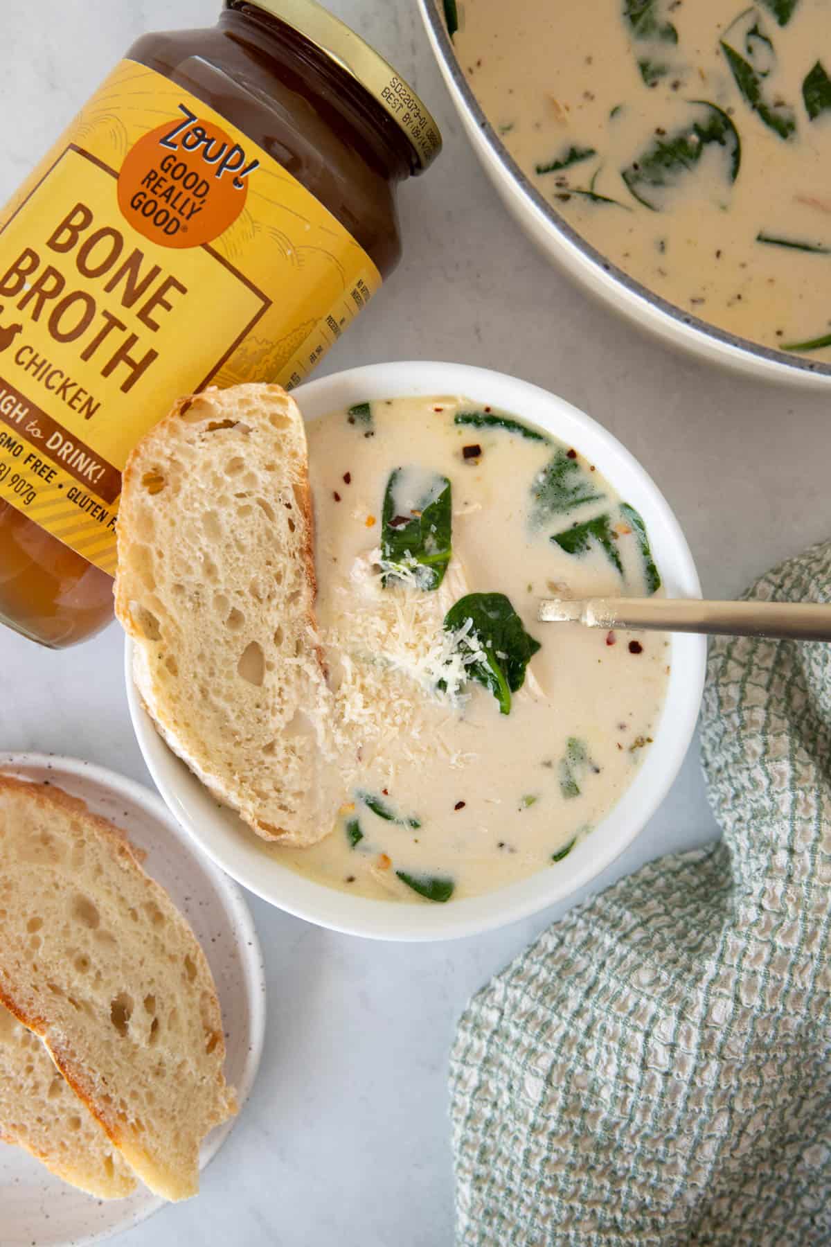 A spoon sits in a bowl of creamy chicken florentine soup with a jar of zoup good real good bone broth on the side.