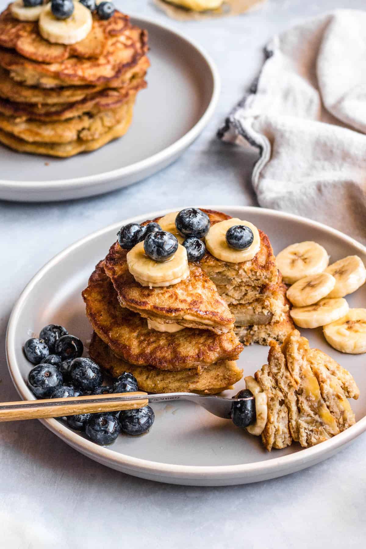 Two plates with stacks of pancakes on top of them and sliced bananas and blueberries.