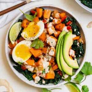 Sweet potato breakfast bowl layered over kale and topped with sliced avocado, boiled eggs, cilantro, lime wedge and a drizzle of tahini.